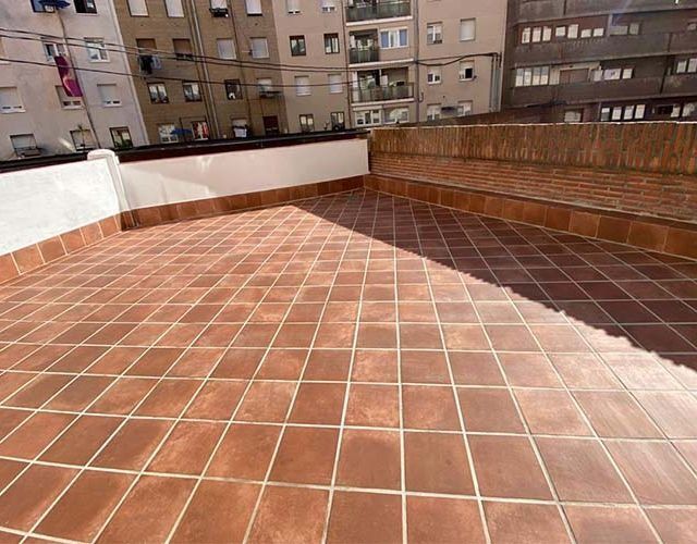 Una terraza soleada con suelo de baldosas de color marrón rojizo, un muro bajo blanco y un banco de ladrillo, situada frente a un apartamento urbano.