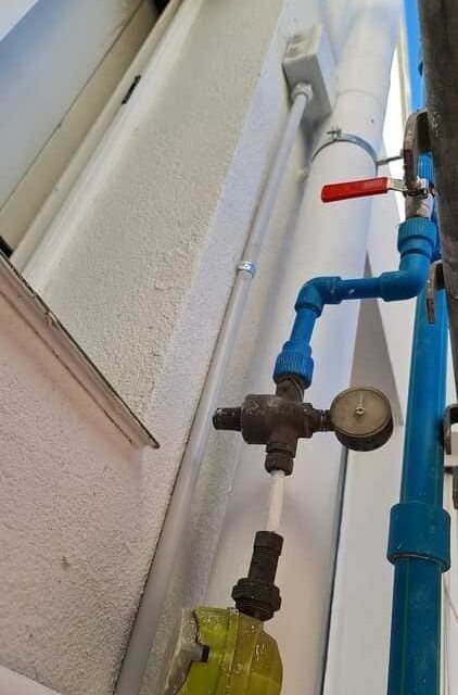 Vista desde un ángulo bajo de un sistema de tuberías de agua azules en una pared exterior, con una válvula de manija roja y un manómetro redondo.