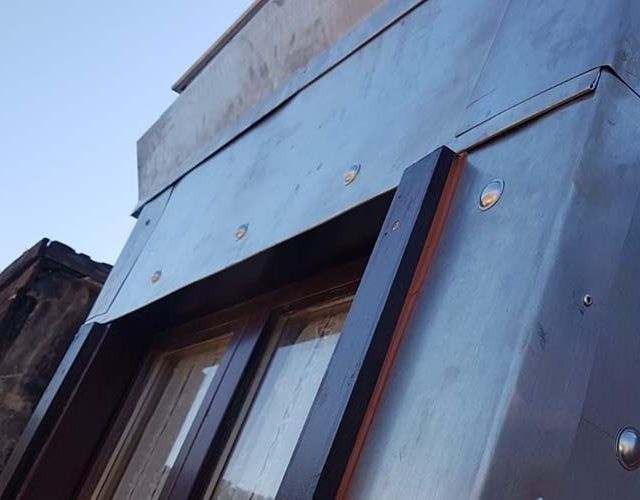 Primer plano de un marco de ventana rodeado de paneles de revestimiento metálico gris fijados con tornillos plateados visibles.