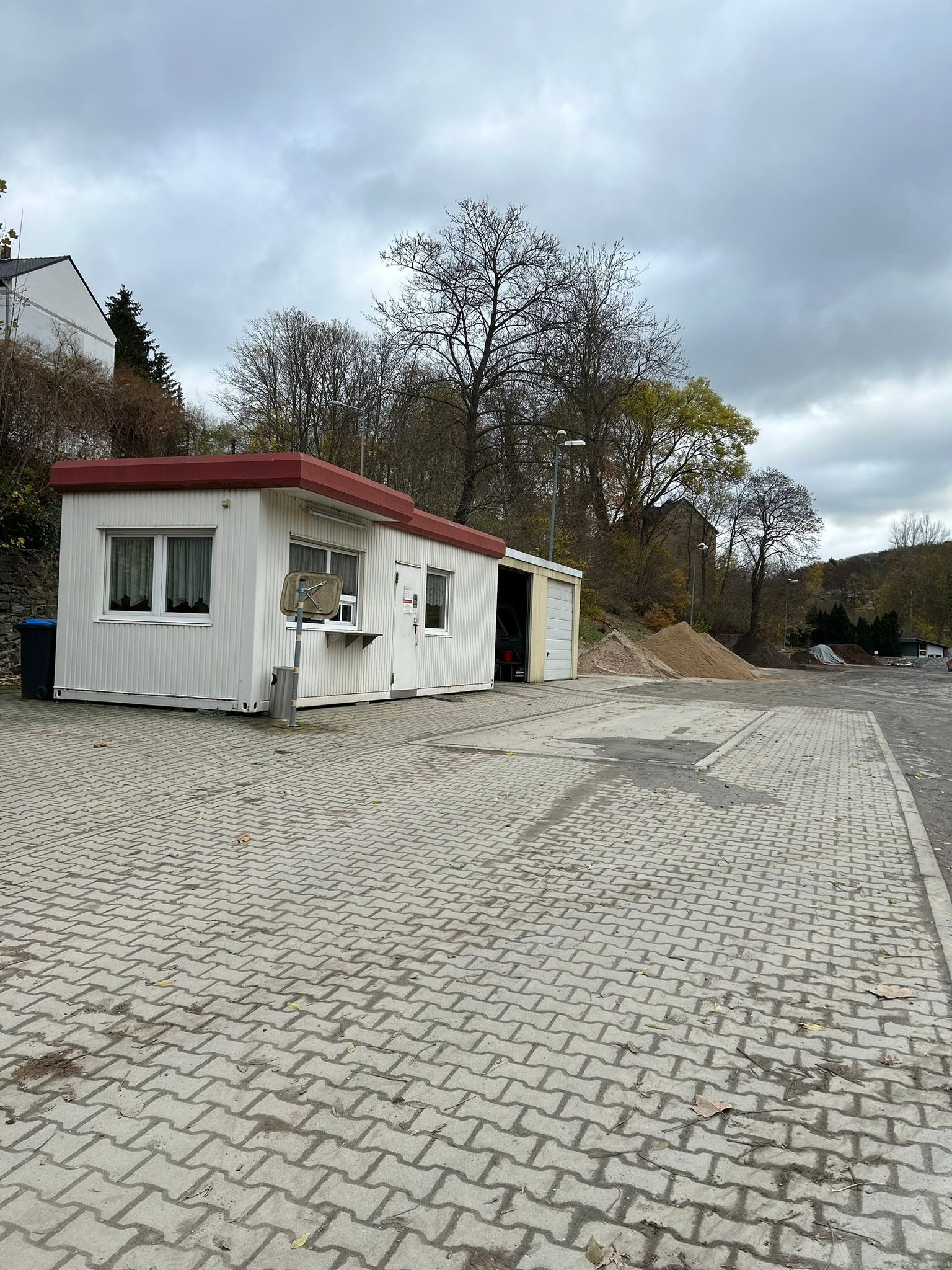 Kleiner weißer Straßenkiosk mit rotem Dach neben einem leeren Kopfsteinpflastergrundstück und Bäumen.