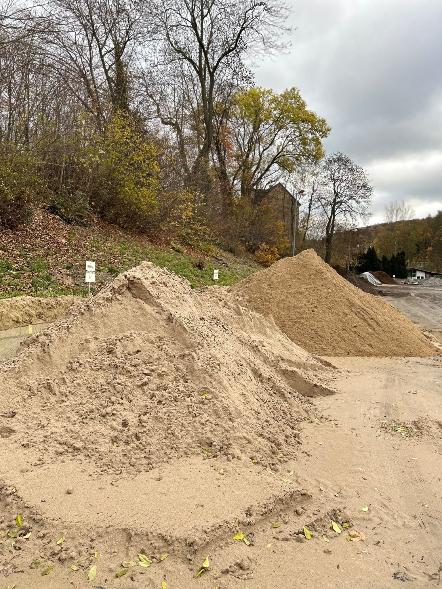 Haufen von Kies und Erde auf einer Baustelle am Straßenrand neben Bäumen und Sträuchern.