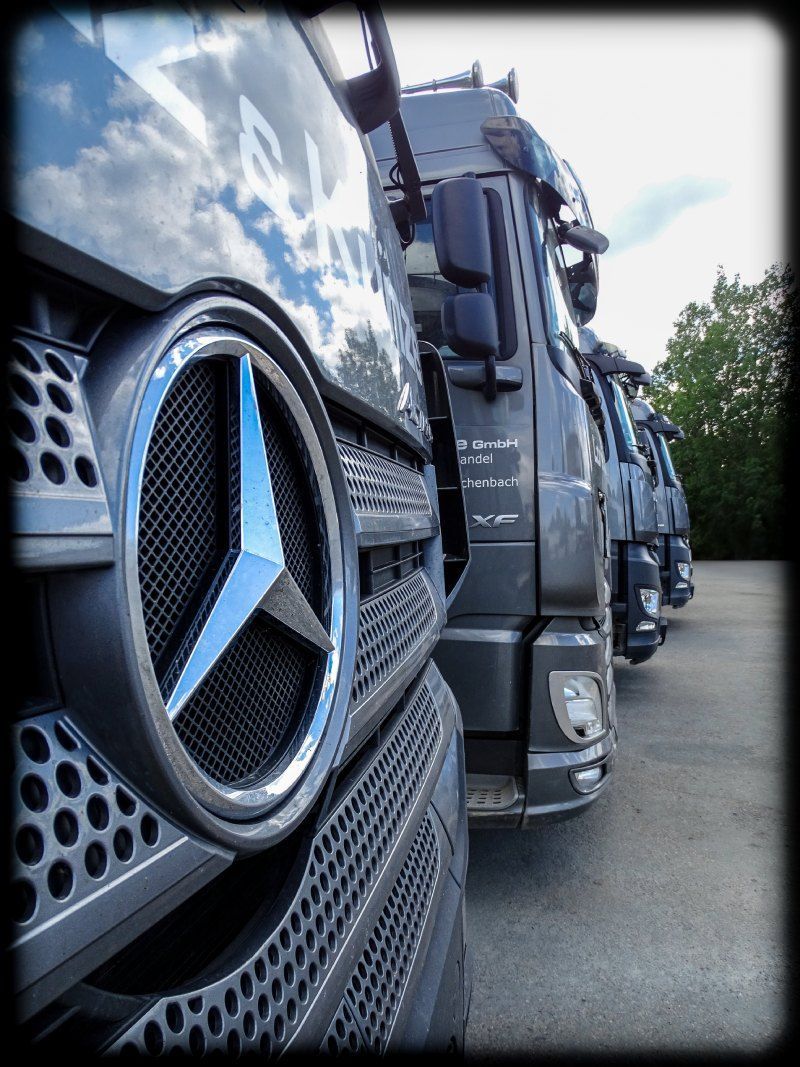 Nahaufnahme des Kühlergrills eines Mercedes-Lkw, dahinter stehen weitere Lkw auf einer Straße.