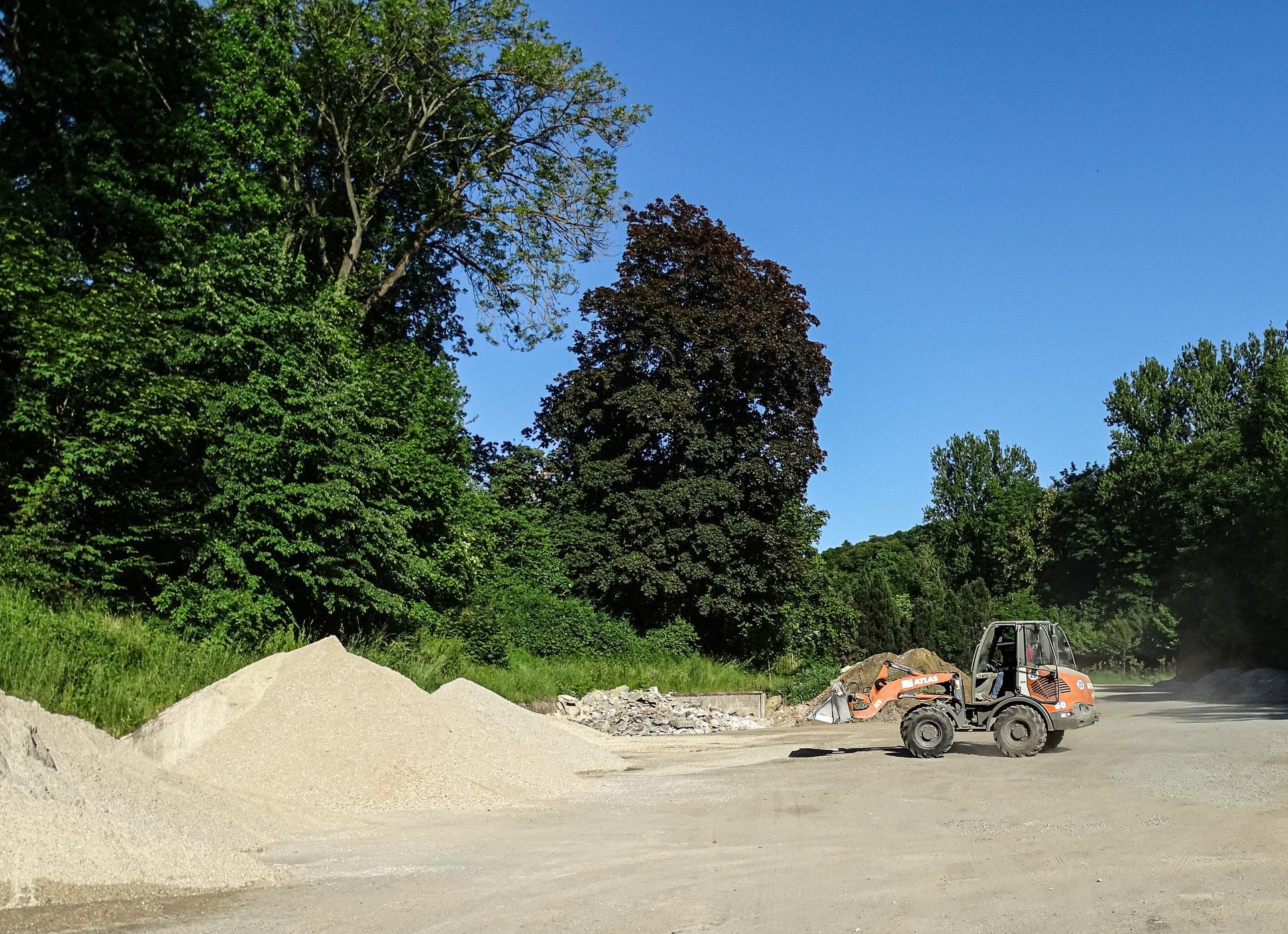 Schotterweg auf einer Baustelle mit Steinhaufen und Bäumen unter einem bewölkten Himmel
