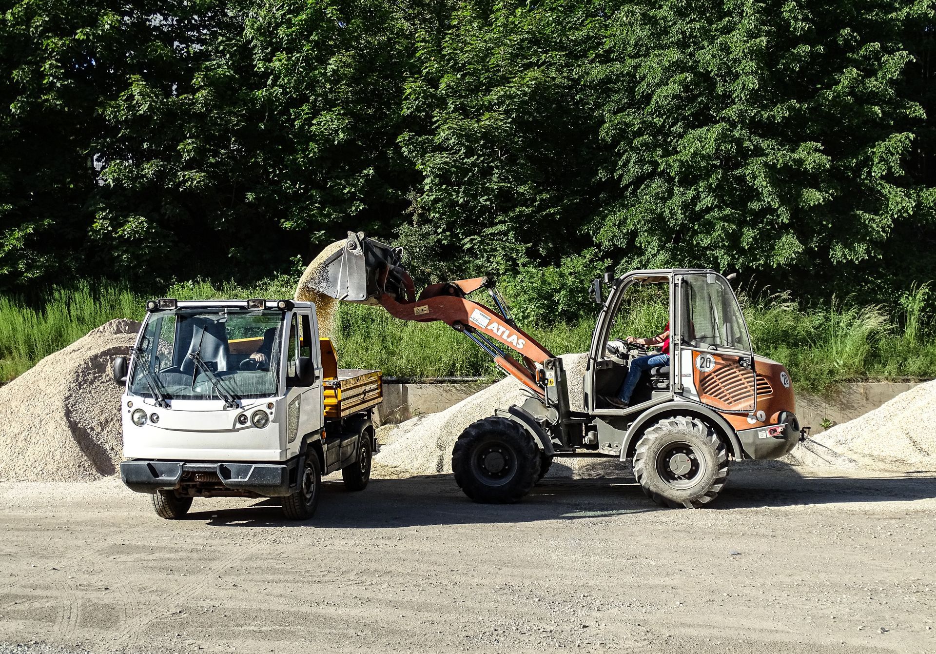 Schotterweg neben einer Baustelle am Hang mit Erdhaufen, Bäumen und bewölktem Himmel