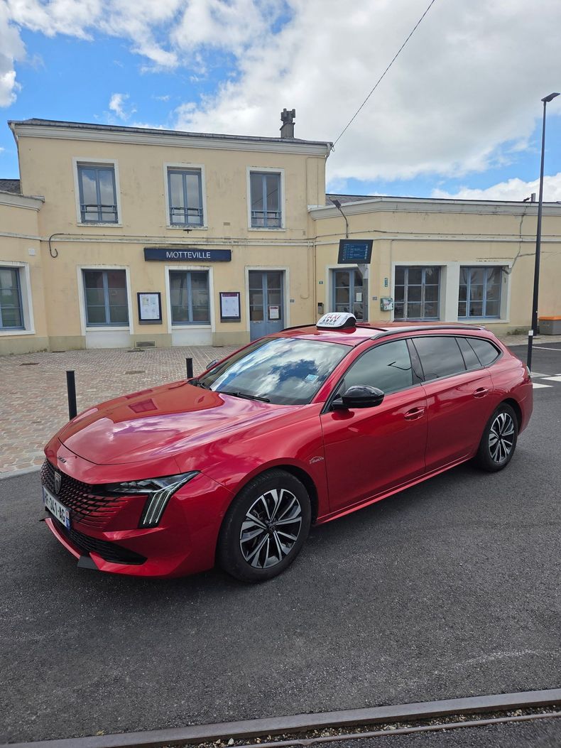 Voiture rouge avec signe taxi, garée dans une rue devant une gare.