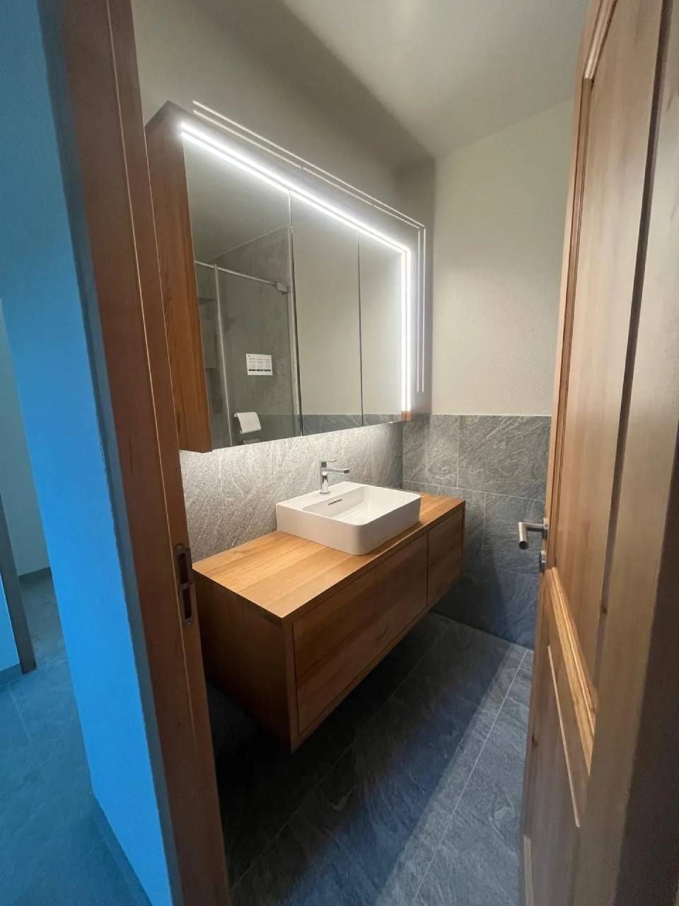 Meuble-lavabo moderne avec armoire en bois, lavabo blanc et miroir éclairé dans une pièce étroite