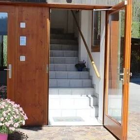 Une porte en bois ouverte laisse apparaître un escalier intérieur, avec une porte vitrée et des plantes en pot à l'extérieur.
