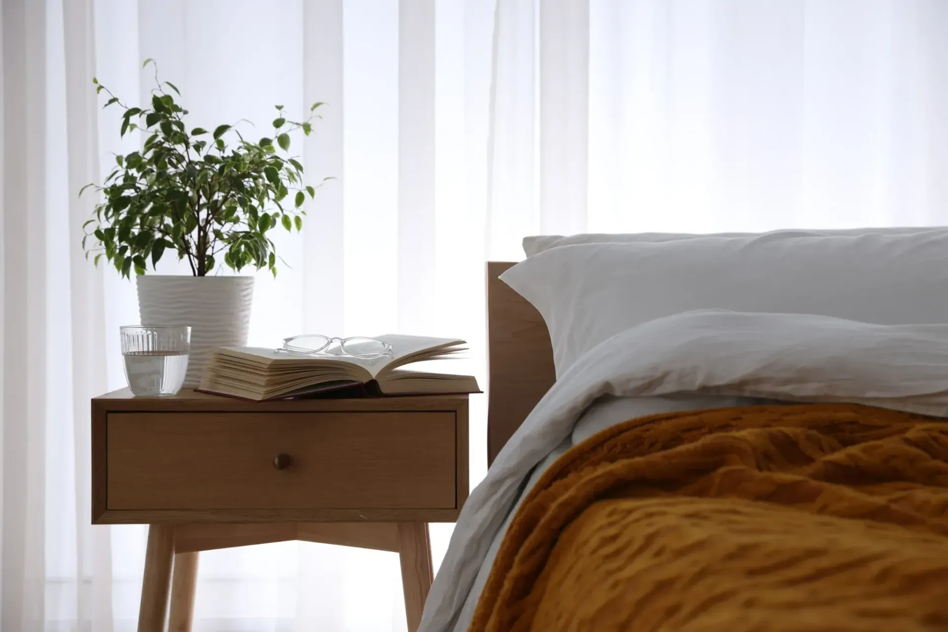 Mesilla de noche de dormitorio con planta, libro y vasos, junto a una cama con manta dorada.