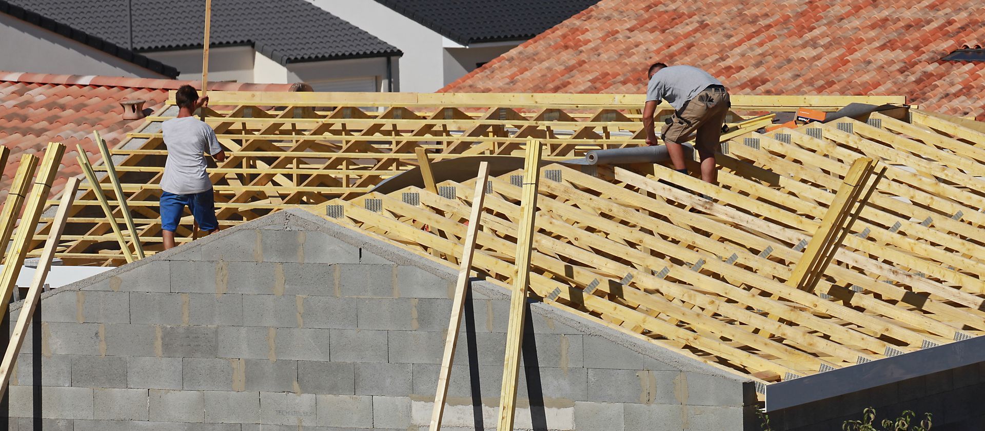 Deux ouvriers du bâtiment sur un toit, en train de construire la charpente. Planches de bois et mur en parpaings gris.