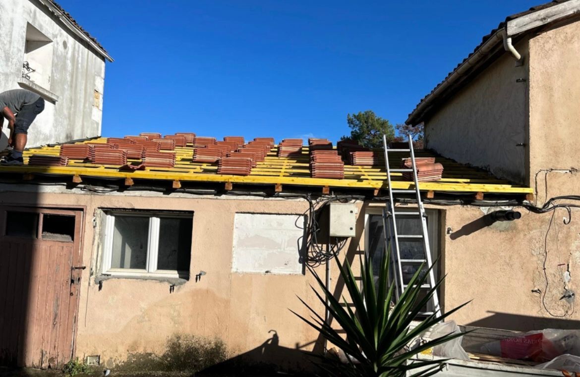 Un homme remplace des tuiles sur le toit. Sous-couche jaune, tuiles orange, échelle. Façade du bâtiment sous un ciel bleu.