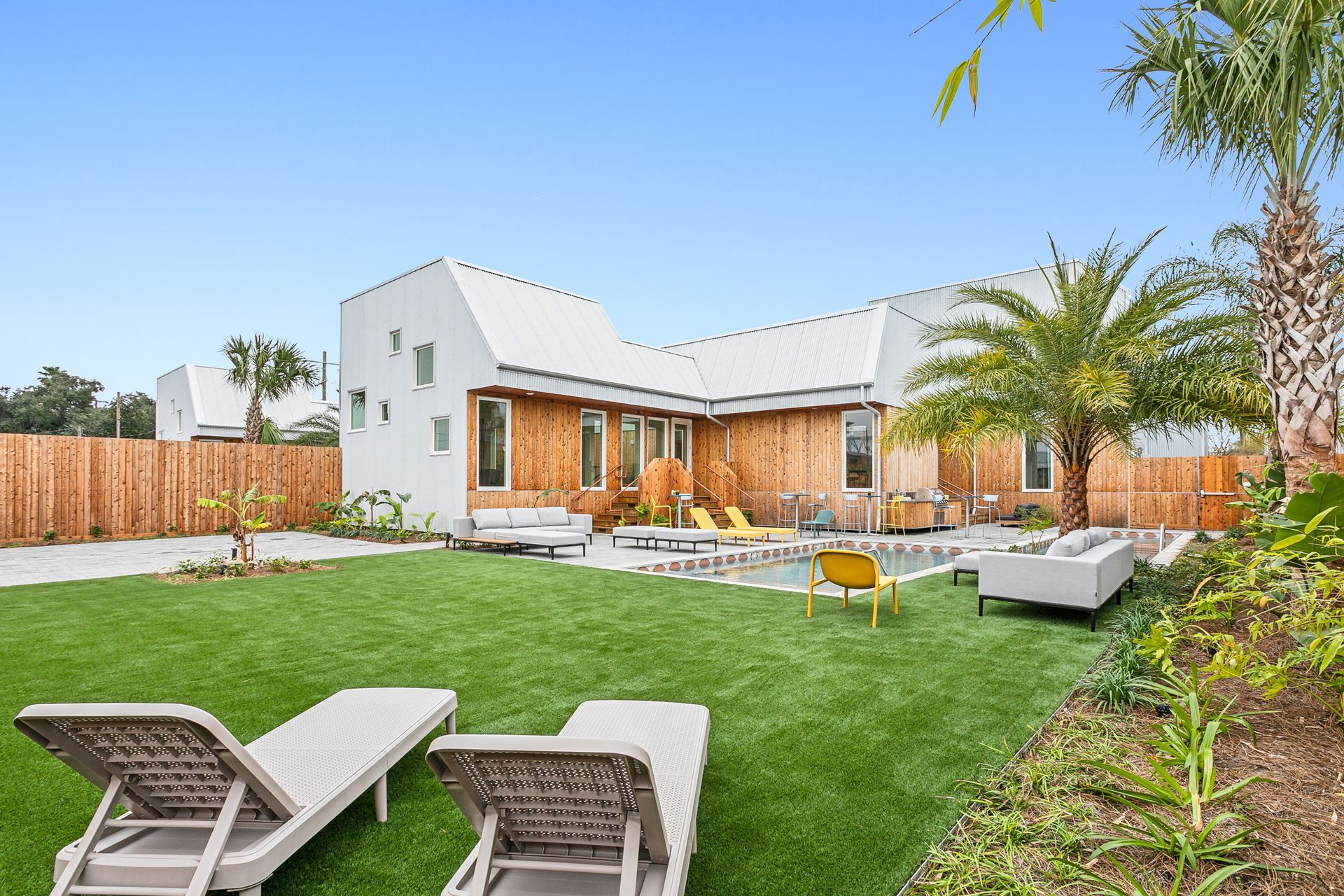 Lawn with lounge chairs, pool, modern house with wooden siding and a palm tree.