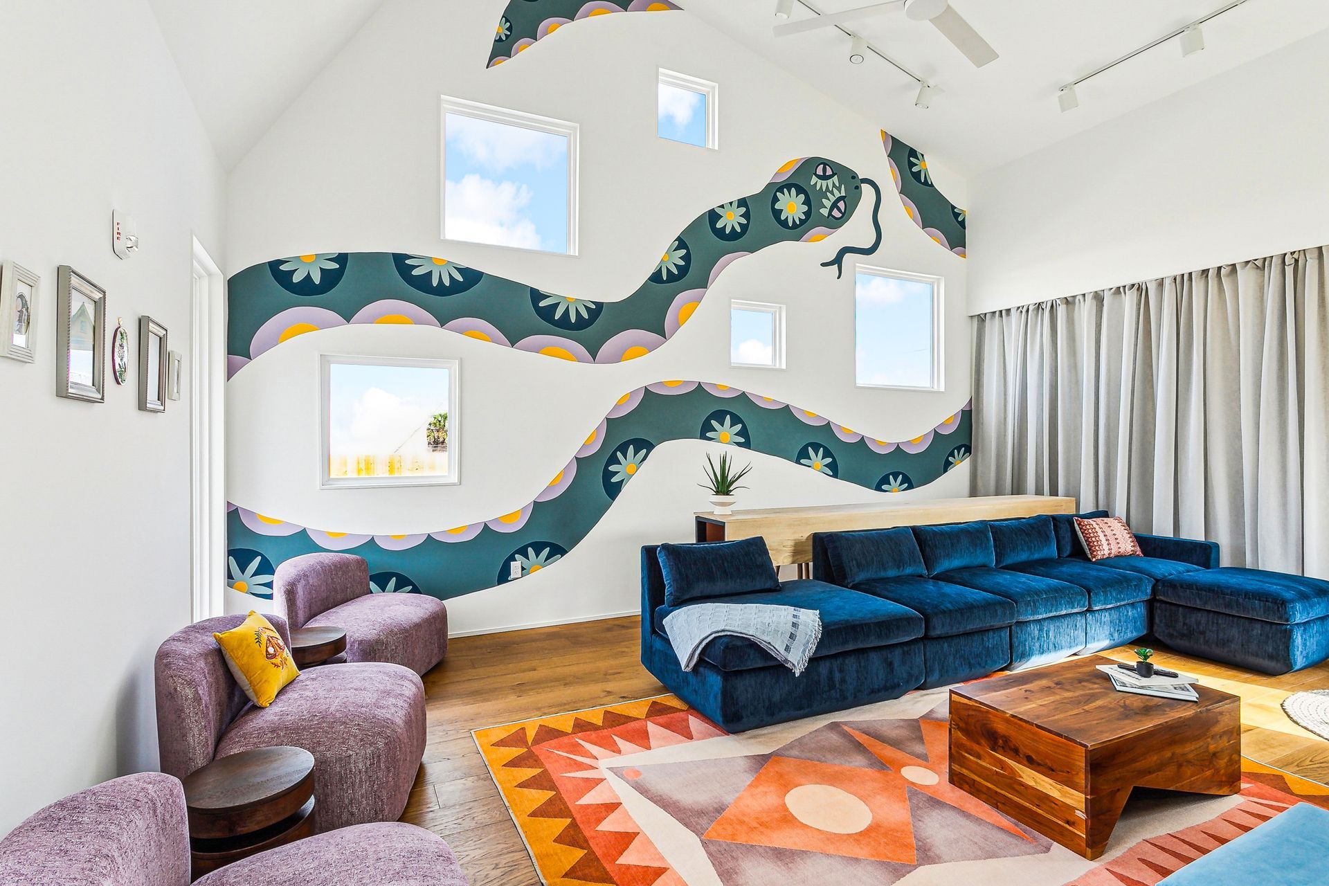 Living room with snake mural, blue couch, colorful rug, purple chairs, windows.