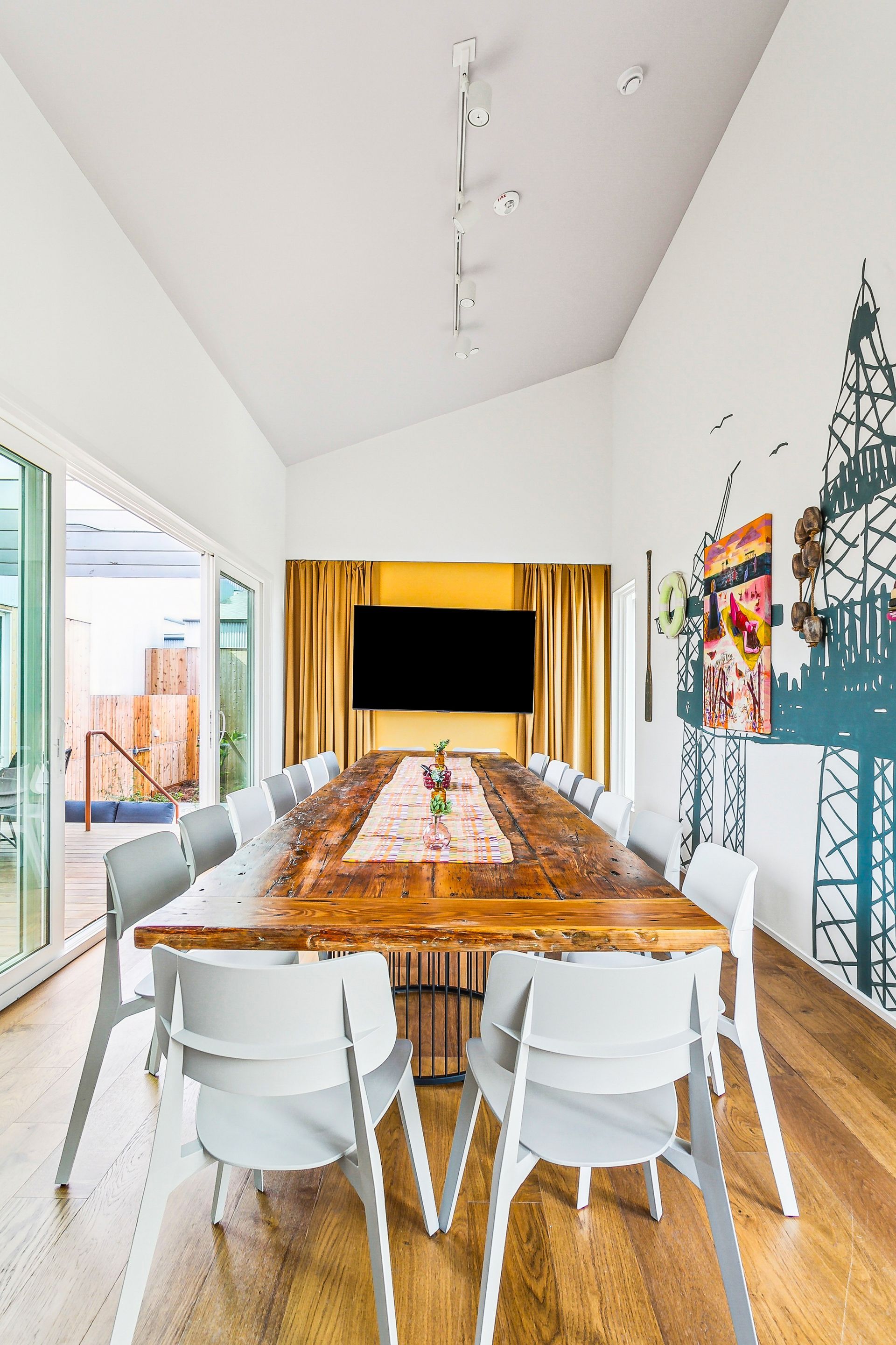 Long wooden table in a conference room with white chairs. Wall art, TV, and window overlooking a pool.