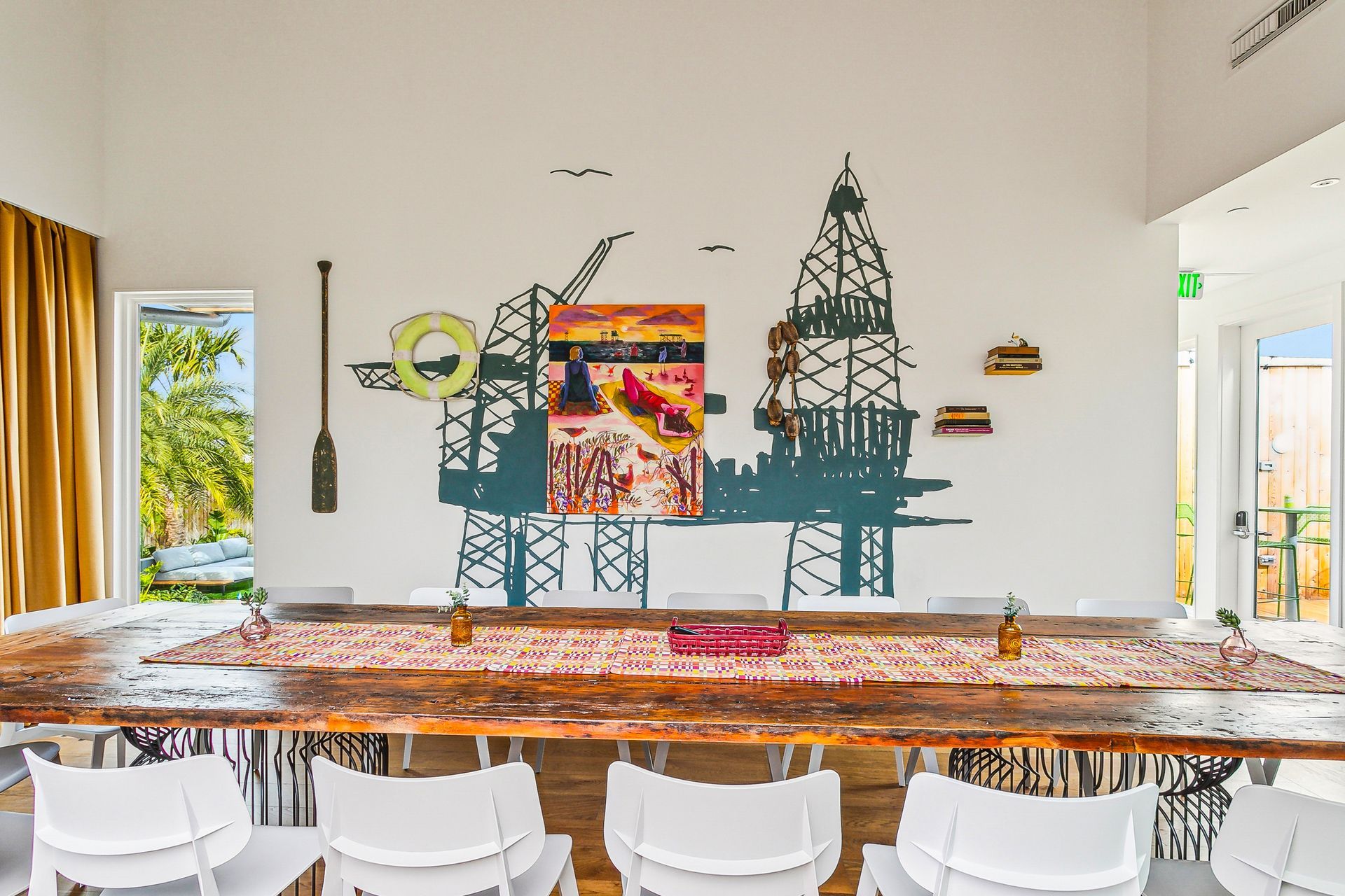 Long wooden dining table with white chairs, art of oil rigs on wall, painting, and gold curtain.