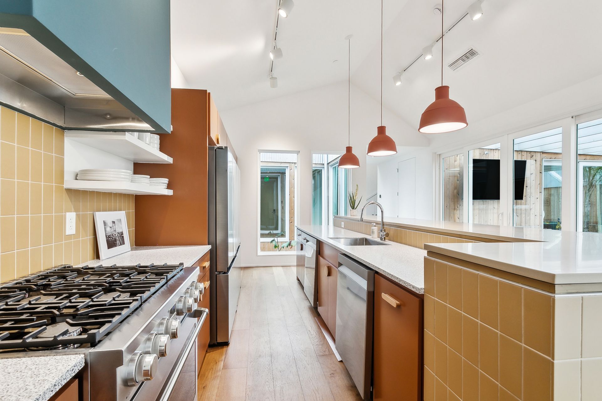 Modern kitchen with orange cabinetry, gold tile, stainless steel appliances, and pink pendant lights.