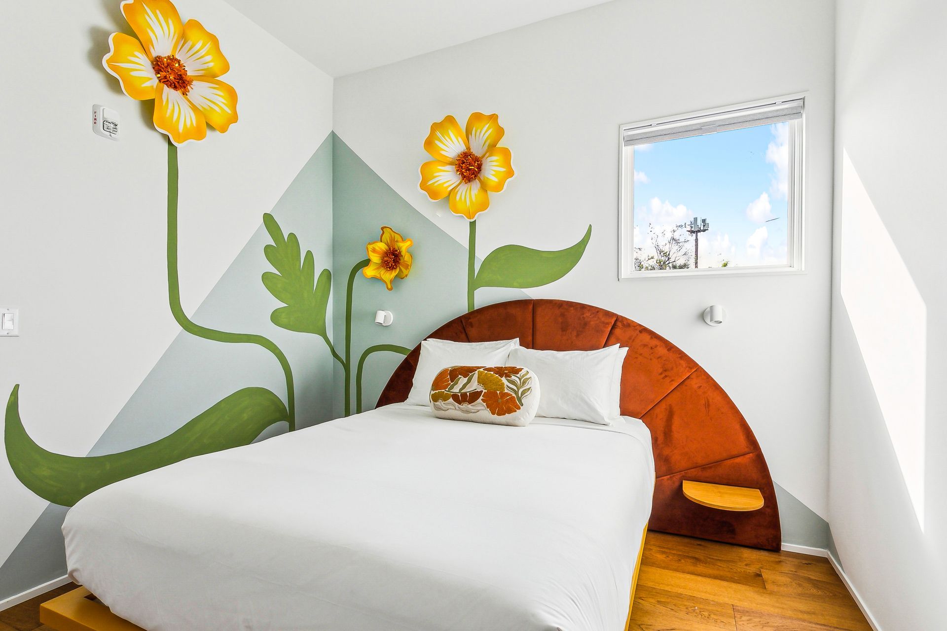 Bedroom with giant painted flowers on the wall, white bedding, wooden floors, and a small window.