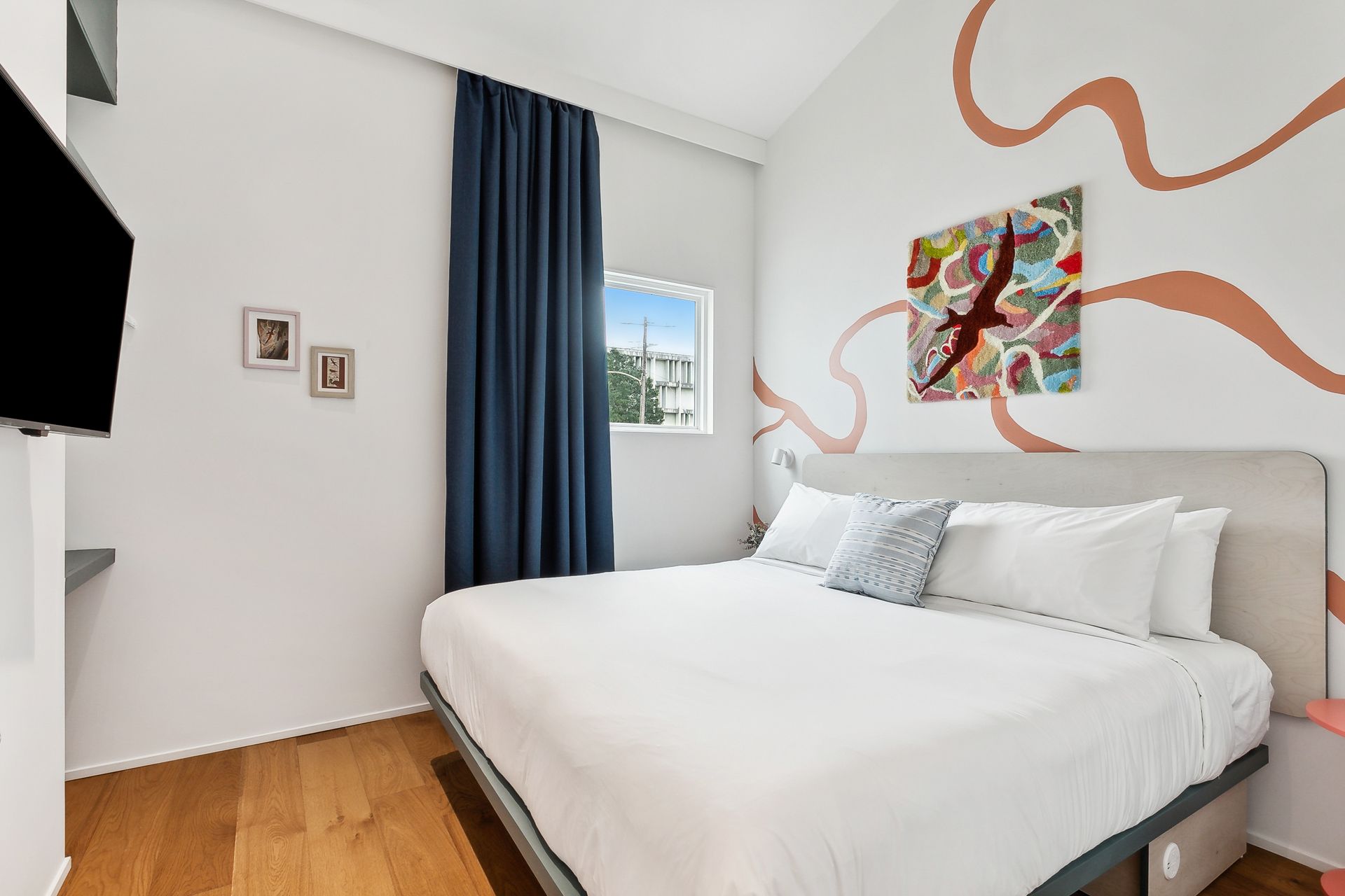 Bedroom with a bed, TV, window, and artwork on a white wall with coral-colored design elements.