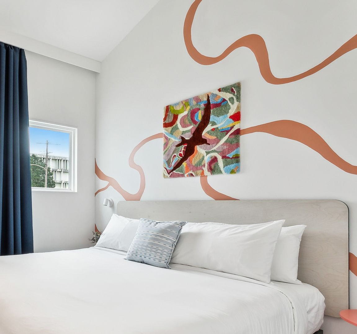 Bedroom with white bedding, art, window, and peach-colored wall design. Blue curtain.