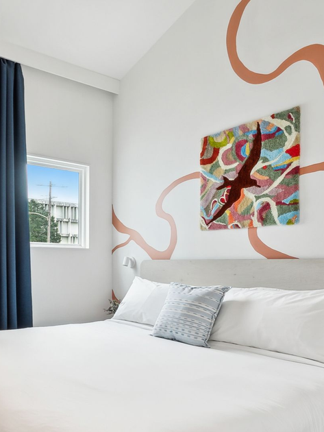Bedroom with white bed, colorful artwork, and window with blue curtain.