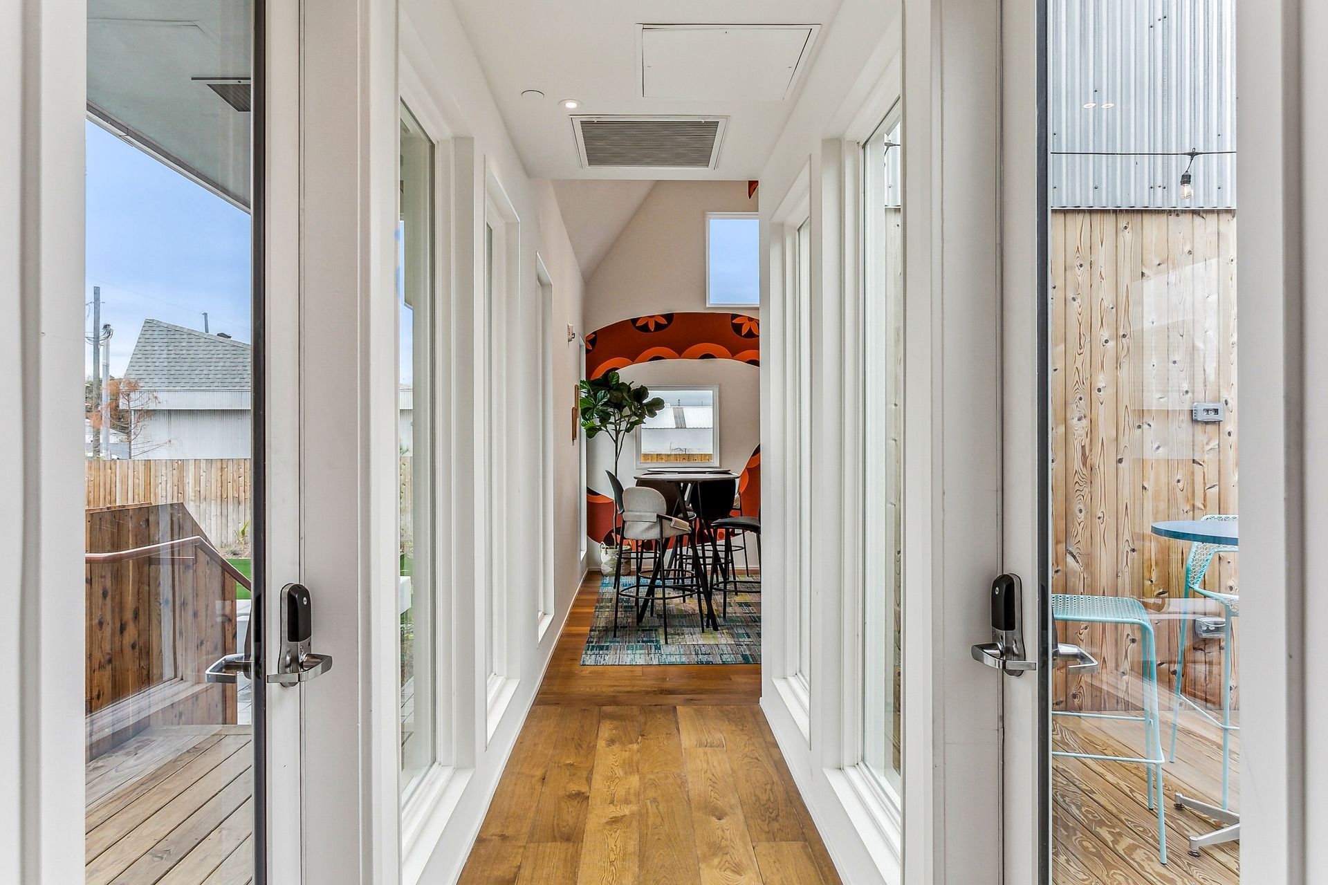 Hallway with wooden floor, framed by glass doors, leading to a dining area with colorful artwork and furniture.