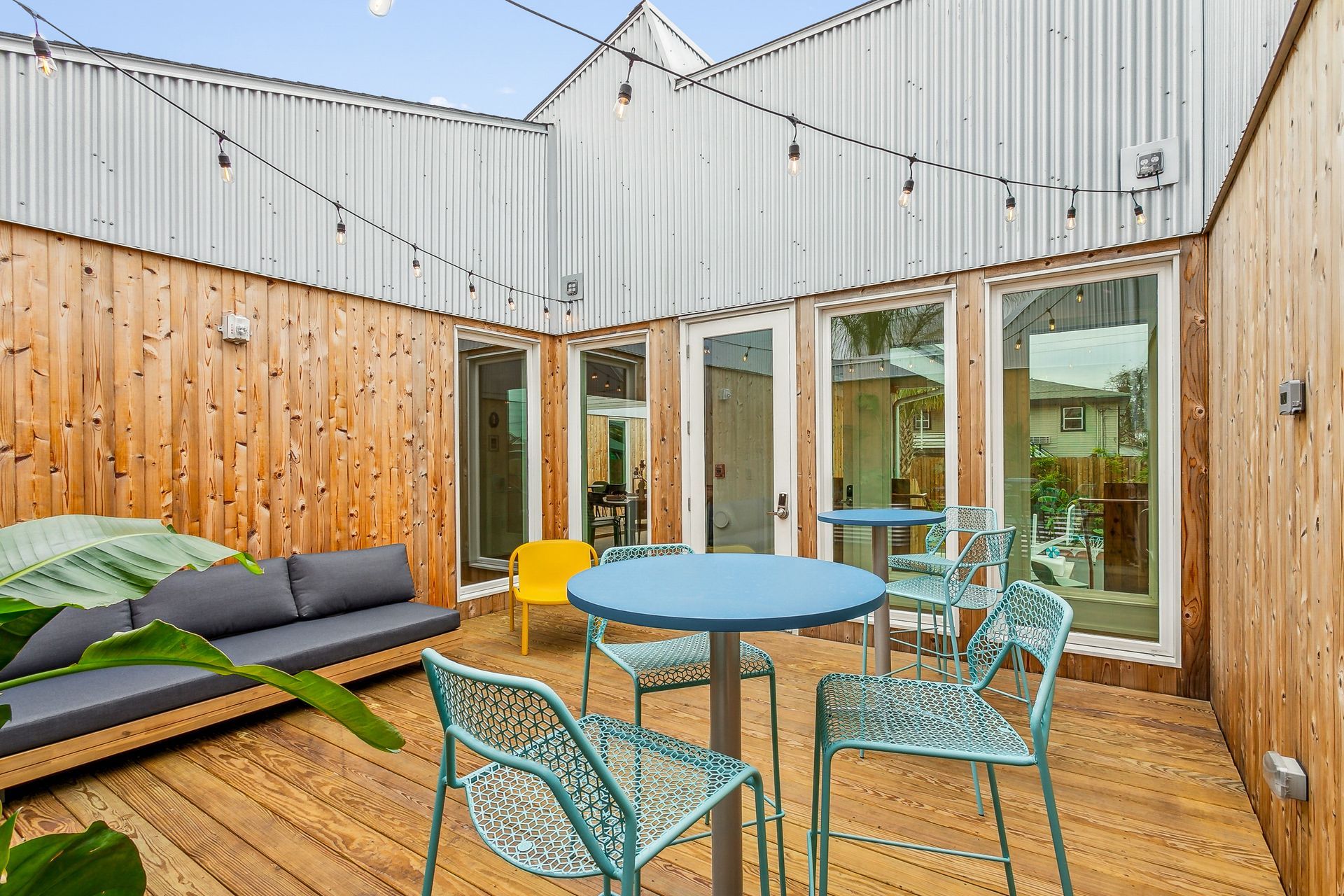 Outdoor patio with blue tables and chairs, wooden deck and walls, and string lights.