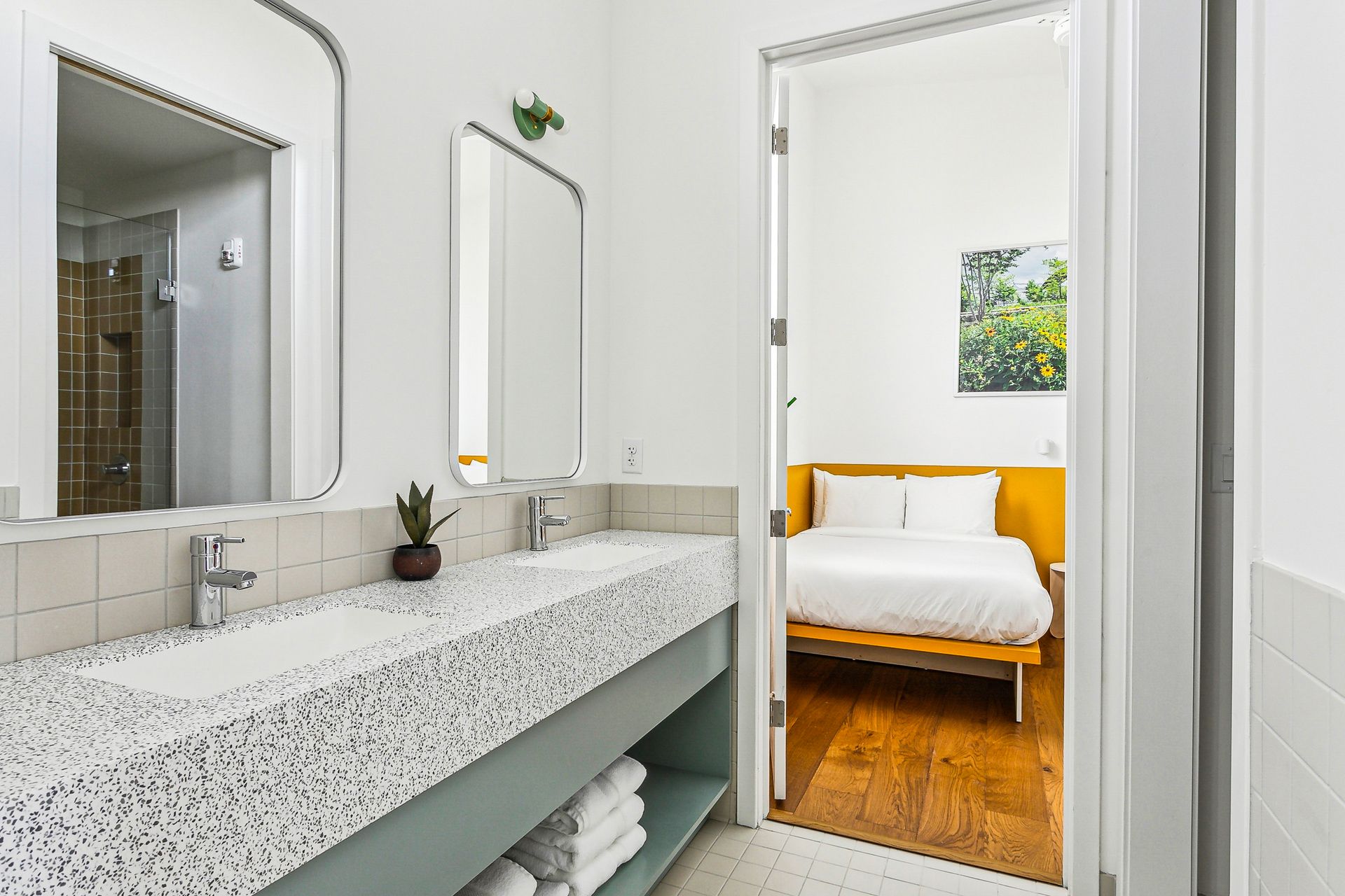 Bathroom with a double sink and a doorway to a bedroom with a bed. Bright, modern design.