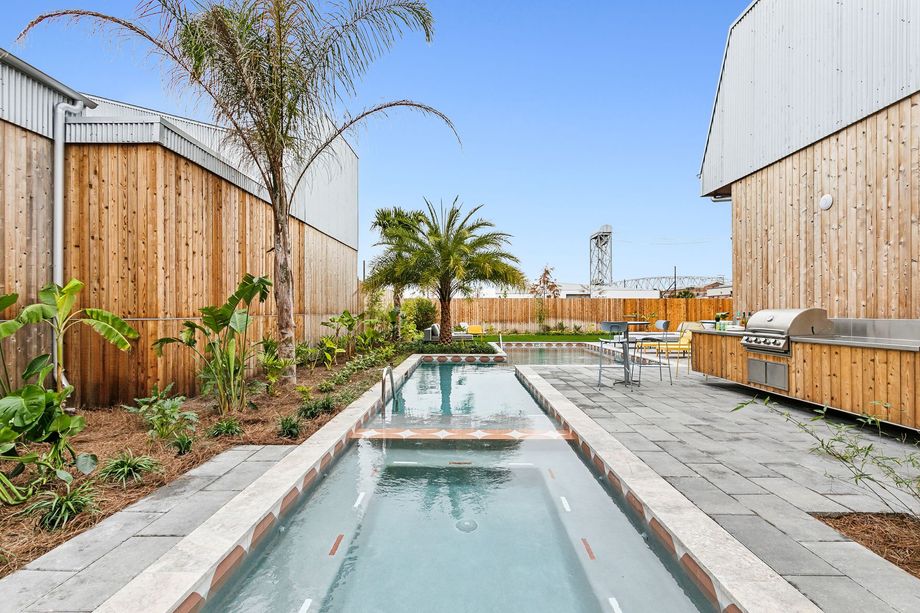 Narrow outdoor pool between two buildings with wood siding, with a grilling area and palms.