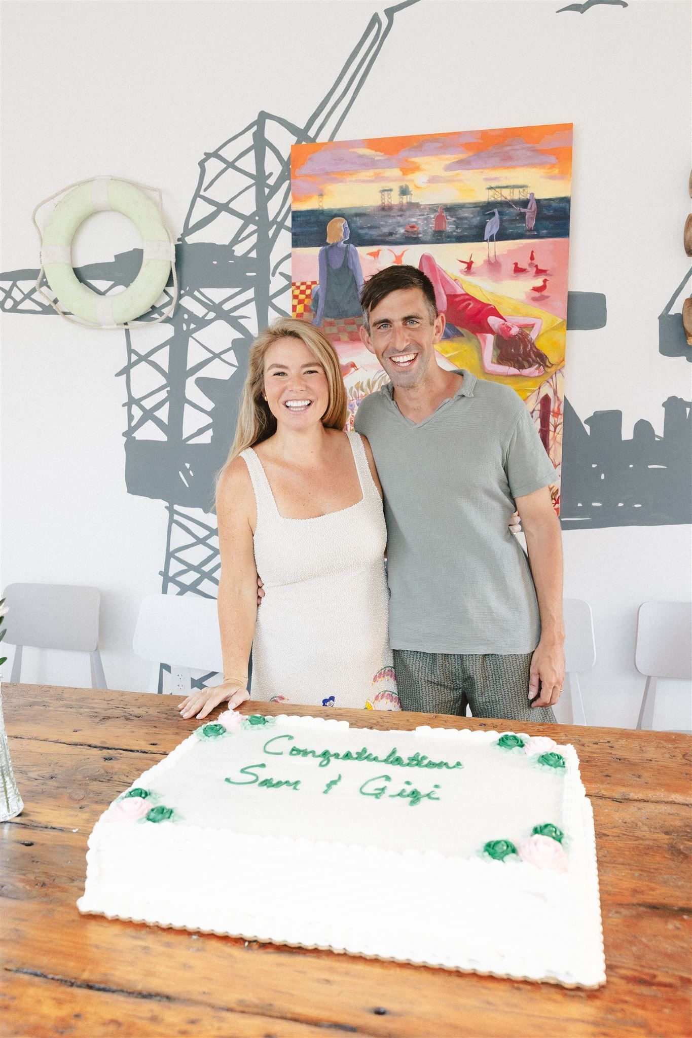 Couple smiling behind a white frosted cake at a party table with colorful wall decorations