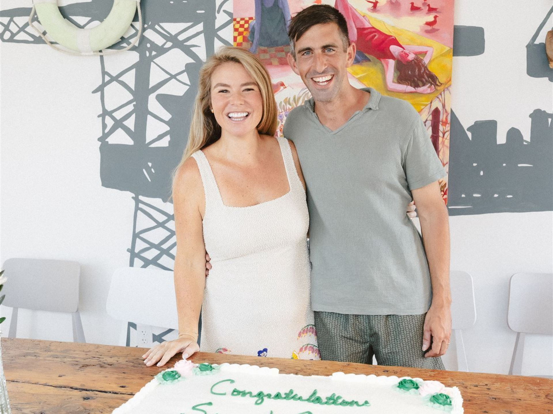 Smiling couple standing behind a table with a congratulatory cake in a bright decorated room