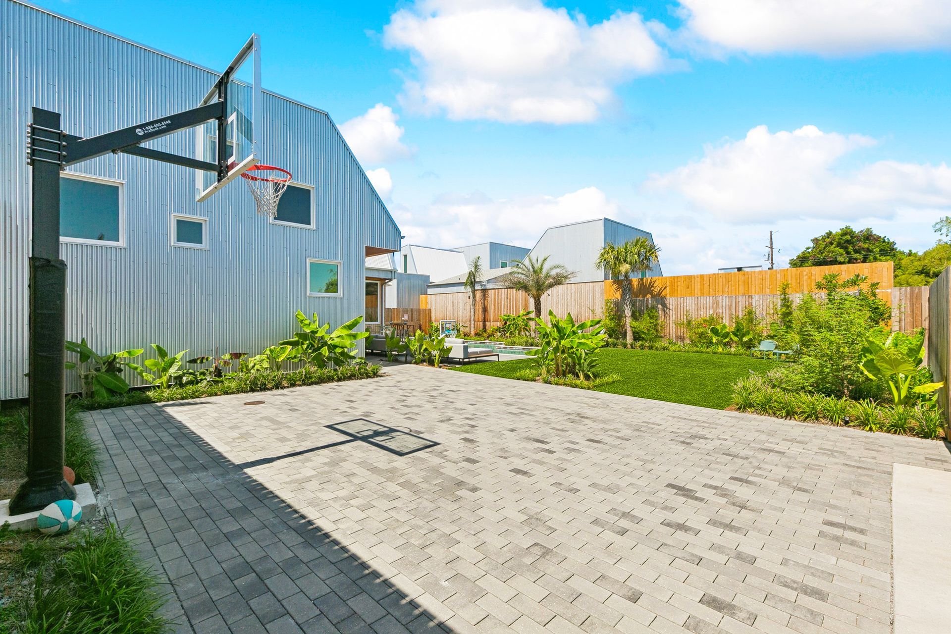 A backyard with a basketball hoop, brick pavement, grass, plants, and a modern silver-sided house on a sunny day.