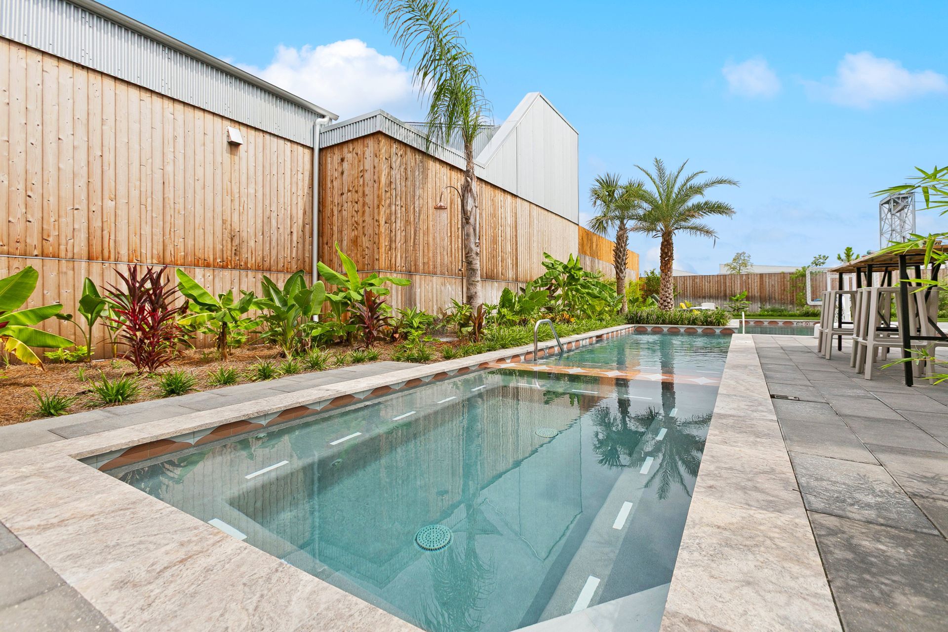 Rectangular pool in a backyard with lush landscaping and wooden walls. Blue sky.