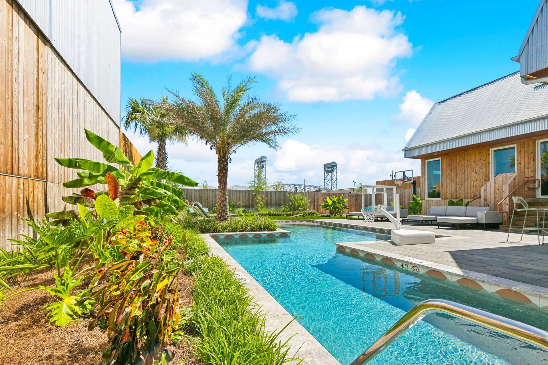 Pool with blue water in a landscaped backyard with palm trees, buildings, and blue sky.