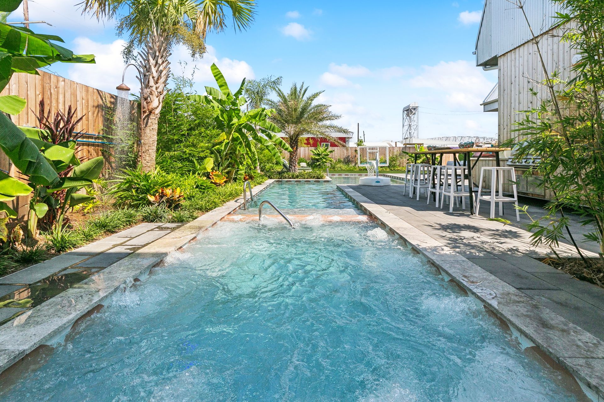 Long rectangular pool with bubbling water, surrounded by lush greenery, near a white building with outdoor seating.
