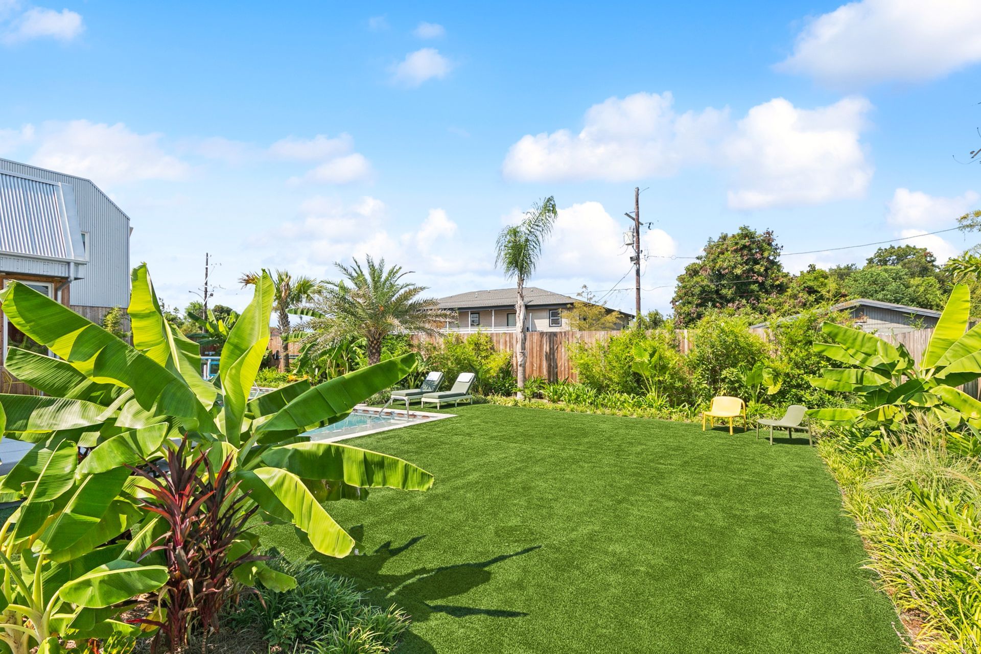 Green lawn in a backyard surrounded by lush plants, with lounge chairs and a blue sky overhead.