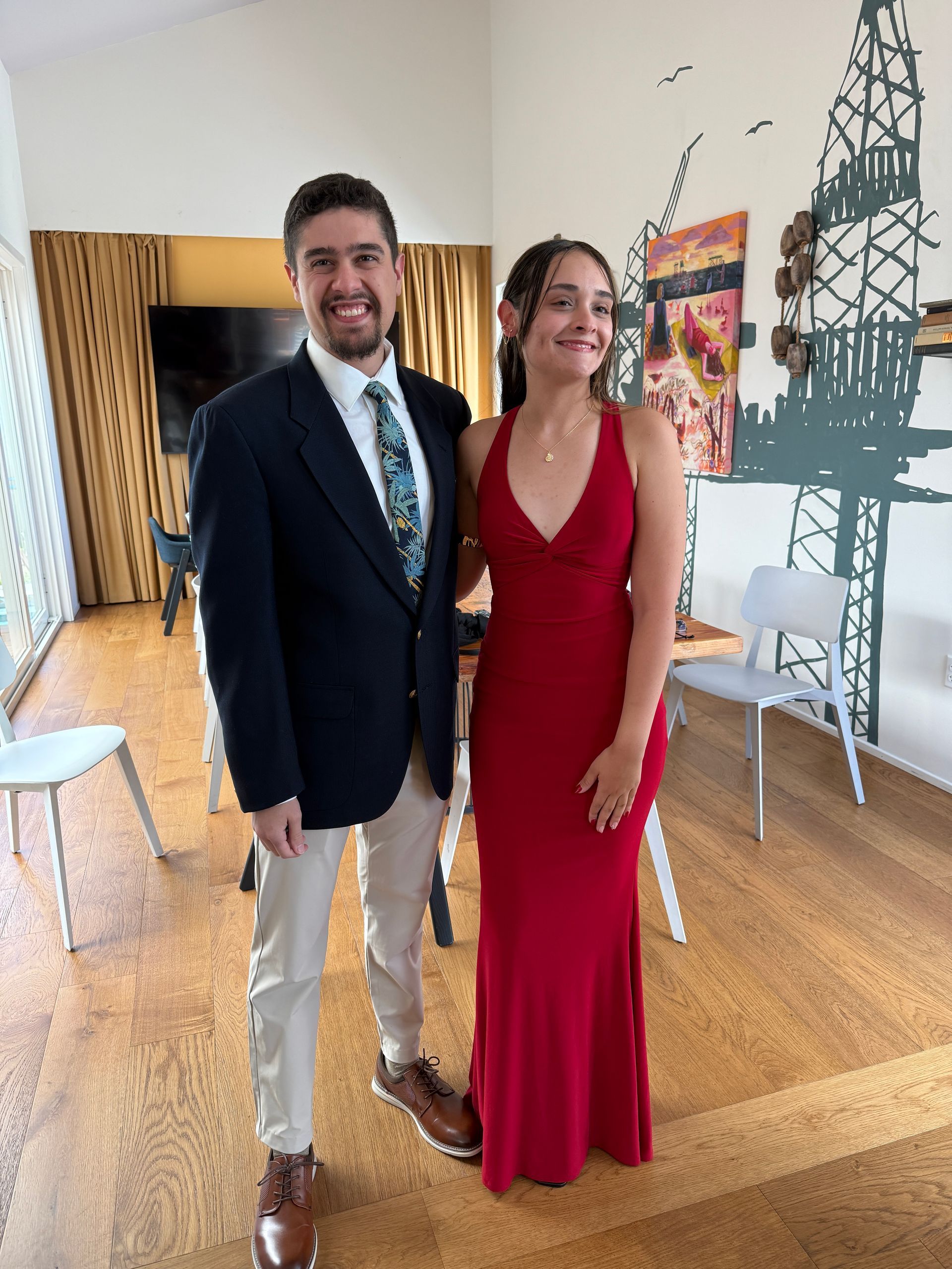 Two people posing indoors, wearing formal attire, one in a navy blazer and the other in a red gown.