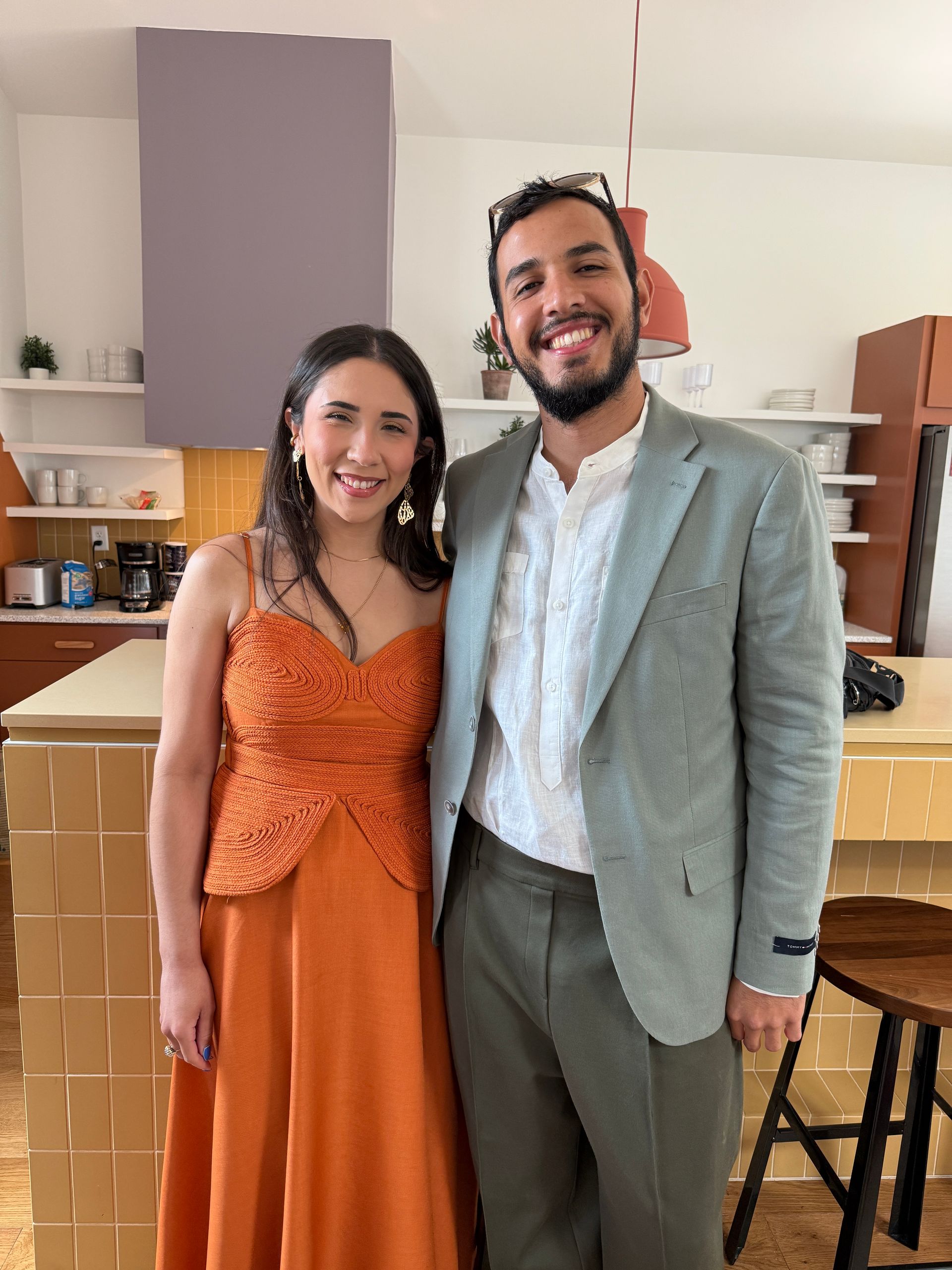 Two people smiling and standing together in a kitchen, wearing orange and light gray outfits.