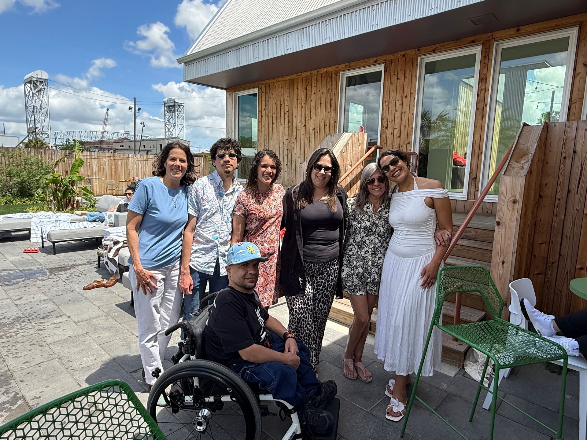 Group of people posing outdoors beside a wheelchair outside a wooden building on a sunny day