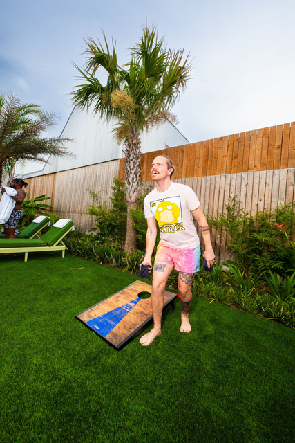 Person standing on a lawn next to a cornhole board, with a palm tree and fence in the background