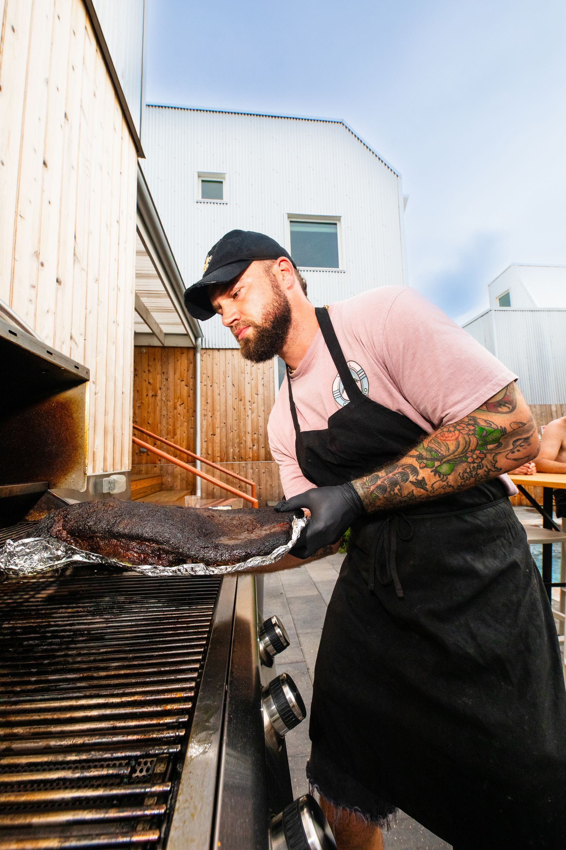 Man with tattoos wearing black apron grilling meat outdoors.