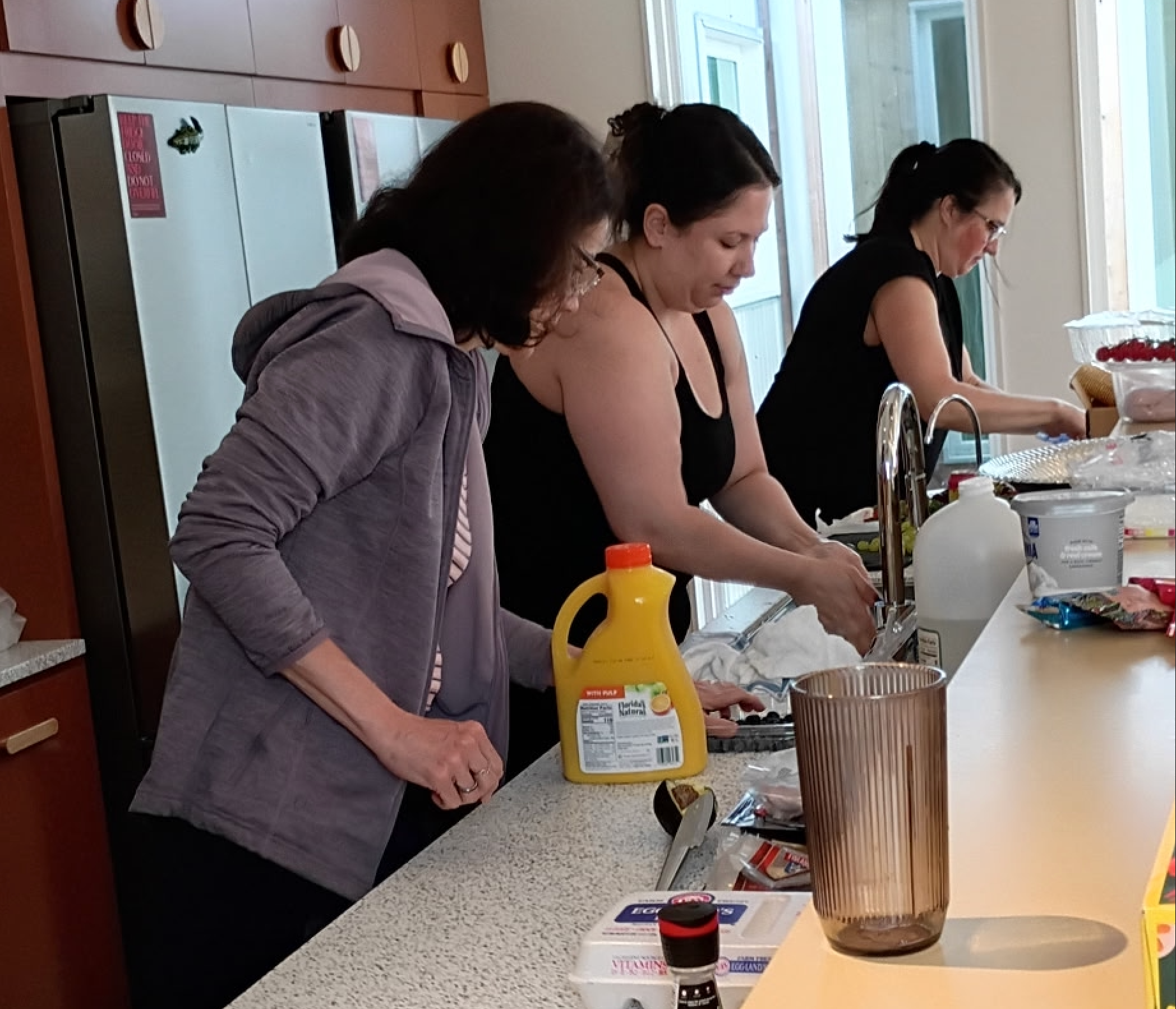 Three people preparing food together at a kitchen counter with bottles and dishes.