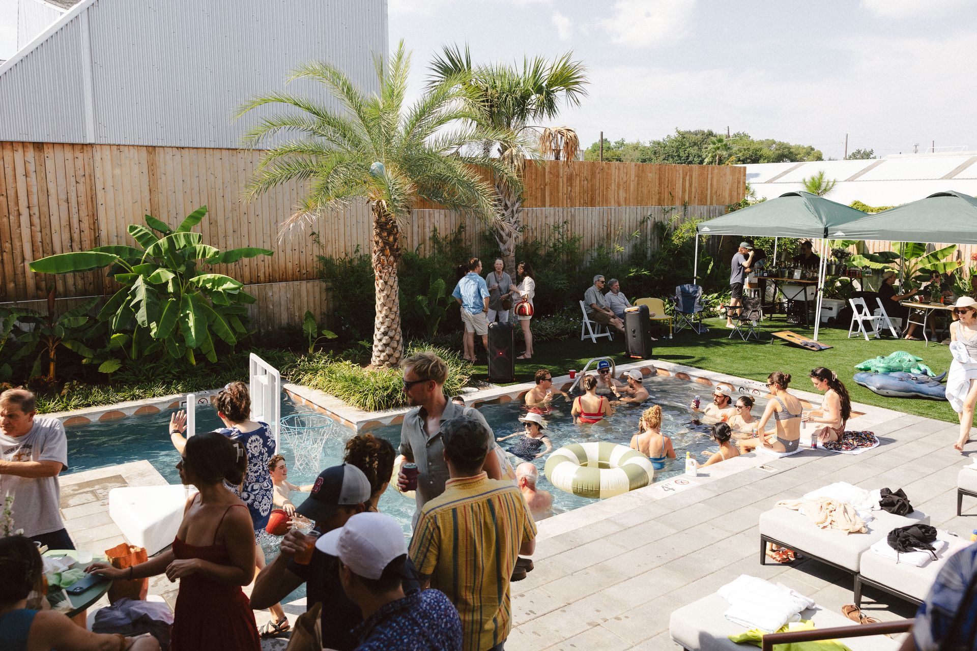 Backyard pool party with people chatting in and around a spa, tables, and a shaded patio.