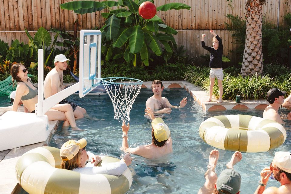 People playing basketball in a pool with inflatable rings. Sunny outdoor setting with a hoop, palm trees, and clear water.