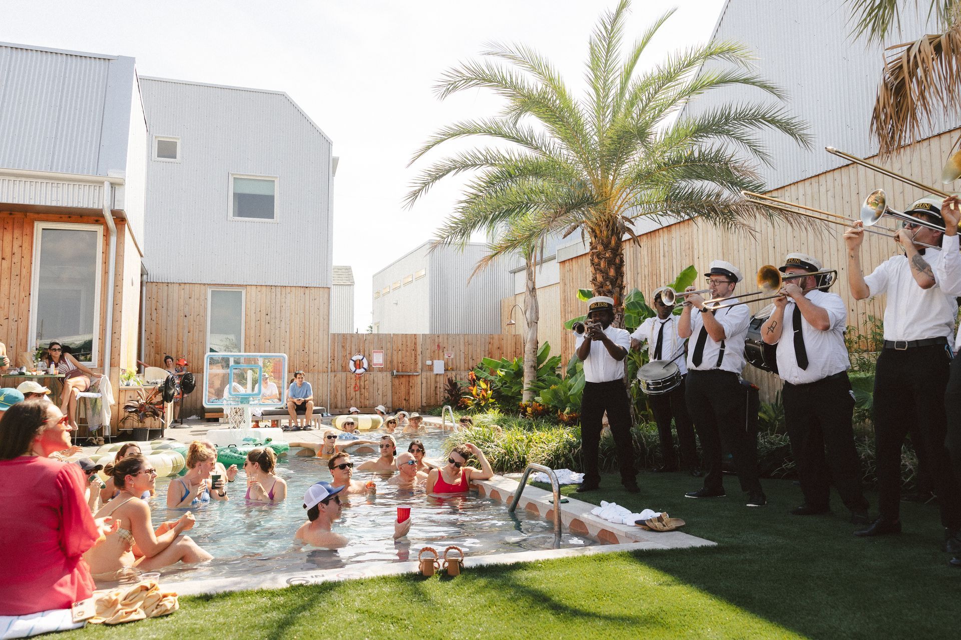 Outdoor gathering by a pool with people mingling and a small band playing near a palm tree.