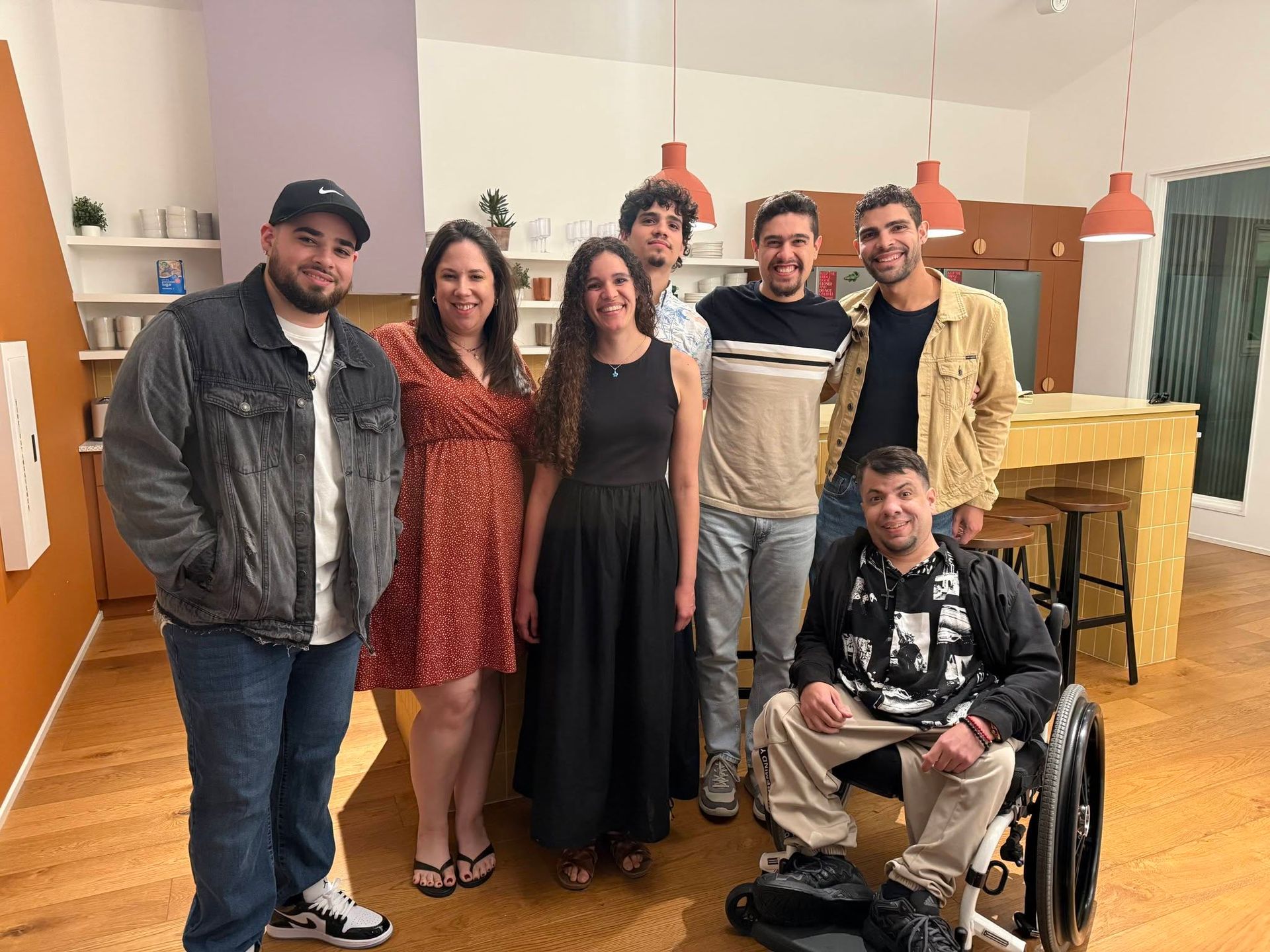 Group of six friends posing together indoors, with one person seated in a wheelchair.