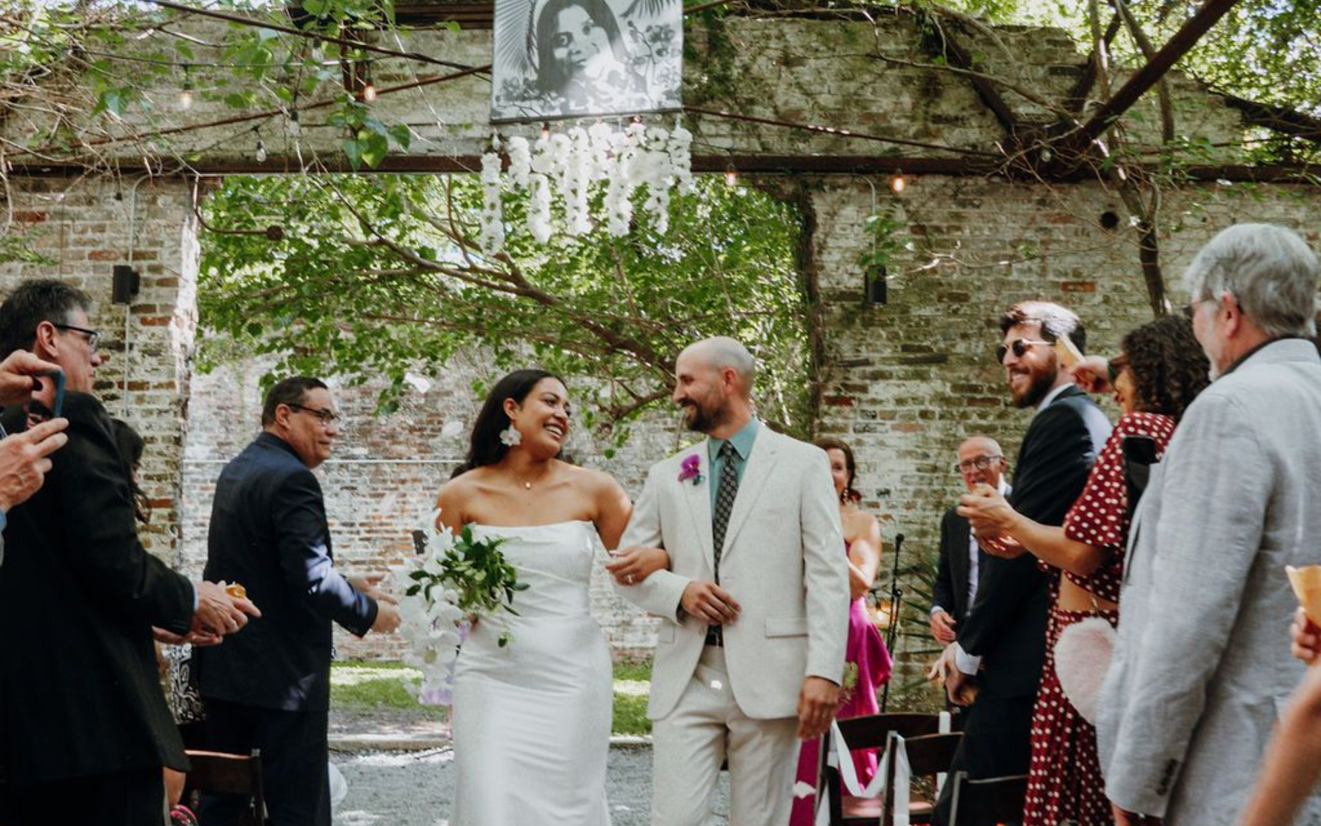 Wedding couple in white outfits smiling during an outdoor ceremony in a rustic garden setting