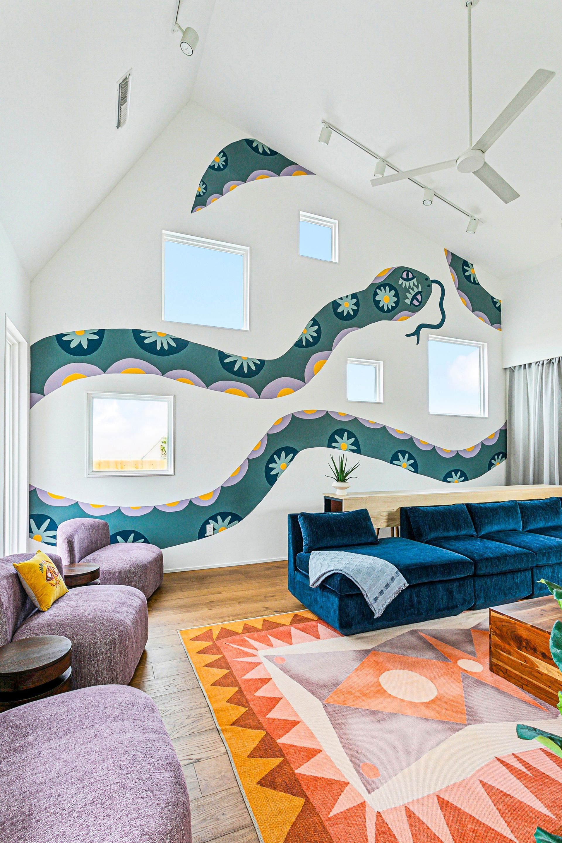 Living room with snake mural on white wall, blue sofa, purple chairs, colorful rug.