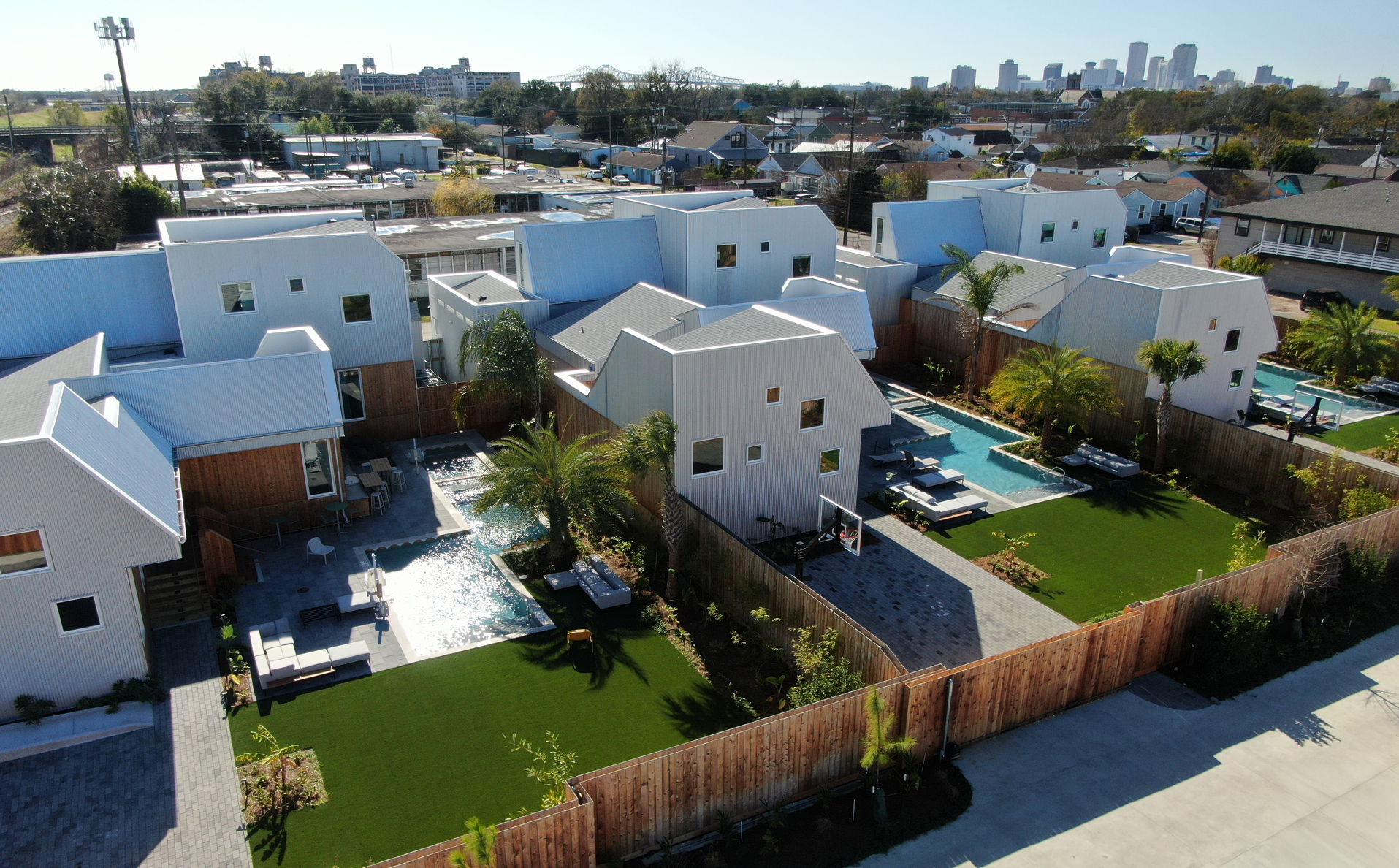 Aerial view of modern white houses with pools, green lawns, and a city skyline in the background.