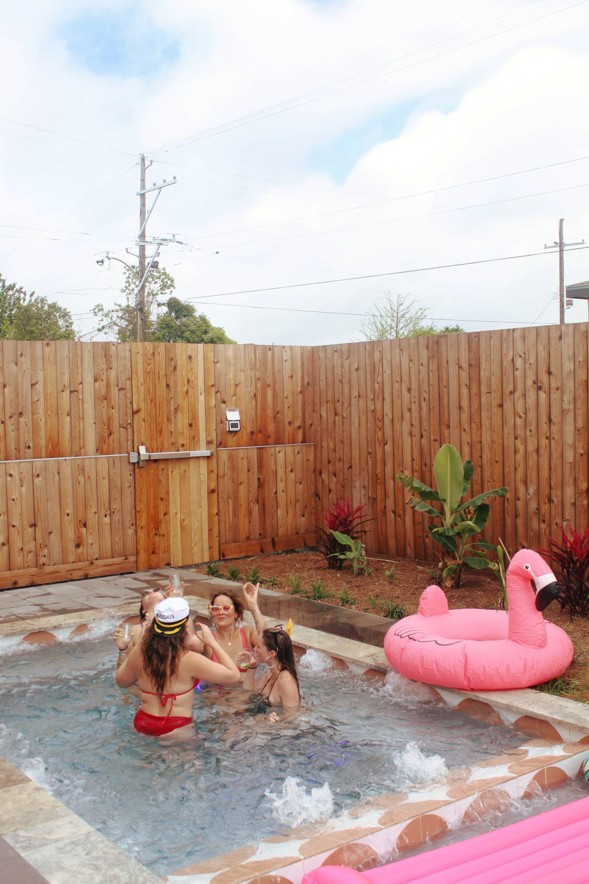 Women in swimsuits enjoy a small pool with a flamingo float in a backyard with a wooden fence.