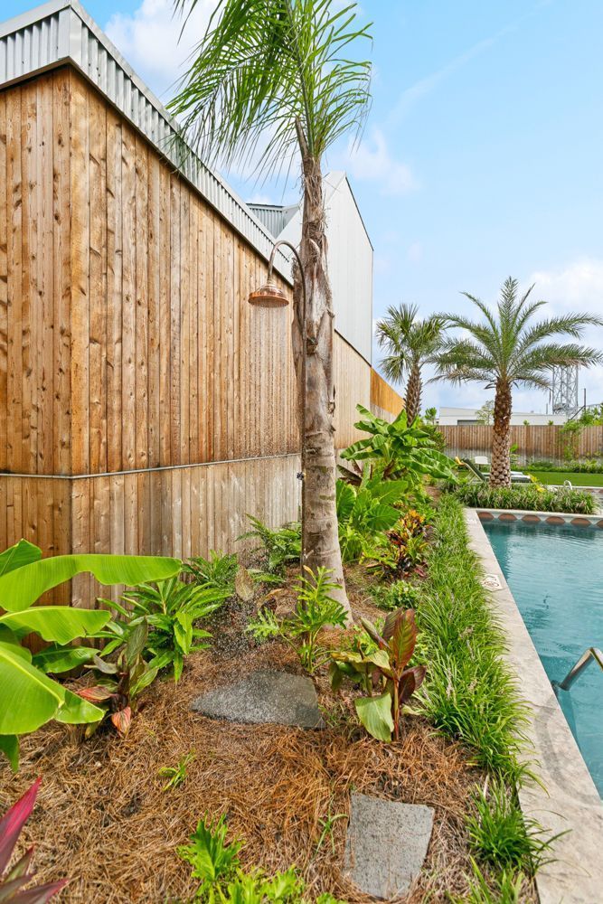 Outdoor shower next to a pool and wooden building with tropical plants and palm trees.