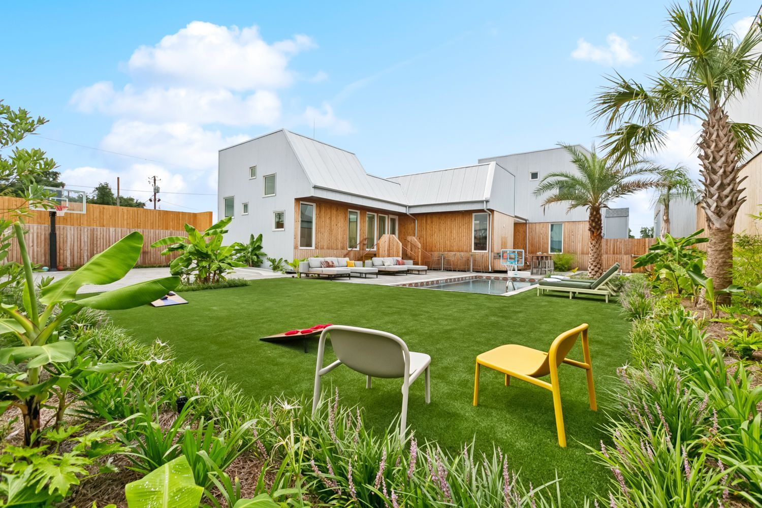 A modern house with a pool and yard, featuring green grass, two chairs, and tropical plants.